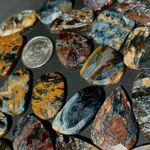 May include: A collection of polished, oval-shaped pietersite gemstones featuring swirling patterns of deep blue, golden yellow, and brown. A silver-colored United States quarter coin is placed among the stones for scale, measuring 0.955 inches in diameter.