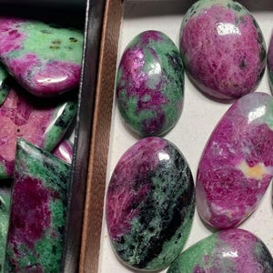 May include: A collection of polished ruby in zoisite cabochons featuring vibrant magenta and deep pink inclusions contrasted against a mottled green and black mineral matrix, displayed in a partitioned container with smooth, rounded surfaces.