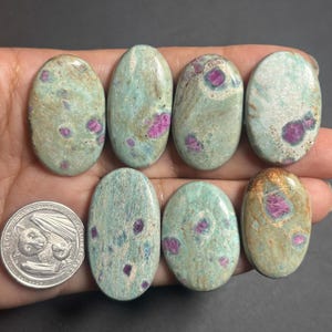 May include: Seven oval-shaped cabochons of green fuchsite with embedded magenta ruby crystals, arranged alongside a silver-colored American quarter coin for scale. Each stone displays a unique pattern of mineral inclusions and polished surfaces.
