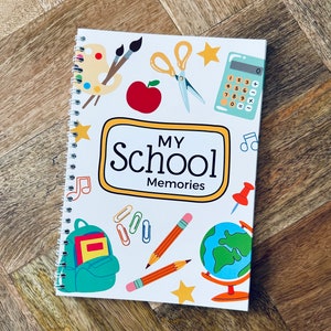 May include: A white spiral bound notebook with a colourful design. The cover features a school theme with illustrations of a paint palette, paintbrushes, a calculator, a globe, a backpack, pencils, and a star. The title of the notebook is "My School Memories".