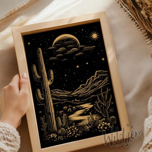 Close-up of hands holding the unframed black and gold desert artwork laid flat on kraft wrapping paper with dried botanical elements.