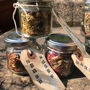 Könnte beinhalten: Vier kleine Glasgefäße mit silbernen Deckeln, gefüllt mit getrockneten Kräutern und Blumen. Jedes Gefäß hat ein braunes Papieretikett, das mit einer Schnur daran befestigt ist. Die Etiketten haben schwarzen Text, der "Sleep", "Peace", "Joy" und "Love" lautet.