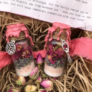 Könnte beinhalten: Zwei kleine Glasflaschen mit rosa Wachssiegeln, gefüllt mit getrockneten Blumen, Kräutern und rosa Himalaya-Salz. Die Flaschen sind mit einem rosa Band zusammengebunden und von getrockneten Rosenknospen umgeben.