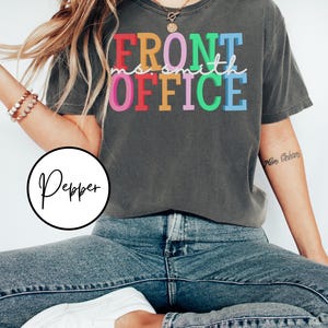 Pu&ograve; includere: Una maglietta grigia scura con il testo "FRONT OFFICE" in colori arcobaleno. Il testo &egrave; in un font di scrittura a mano.