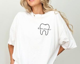 May include: White t-shirt with a black outline of a tooth and the name "emma" written across the tooth.