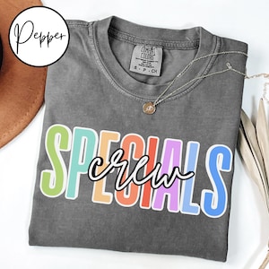 May include: A gray t-shirt with the word "SPECIALS" in colorful block letters and "crew" in a cursive font. A gold necklace with a pendant is on the shirt. The shirt is folded on a white surface.