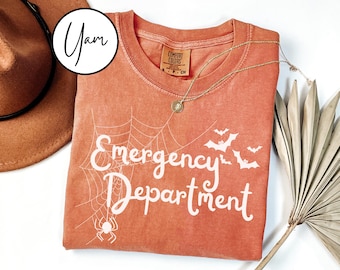 May include: An orange t-shirt with the words "Emergency Department" in white, along with a spider web, spider, and bats. A gold necklace with a pendant is on the shirt. A brown hat is in the upper left corner.