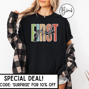 May include: Black t-shirt with the words "FIRST grade" in colorful block letters. The shirt has a crew neck and short sleeves. The text is outlined in white. The shirt is worn with a plaid shirt.
