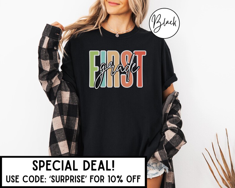 May include: Black t-shirt with the words "FIRST grade" in colorful block letters. The shirt has a crew neck and short sleeves. The text is outlined in white. The shirt is worn with a plaid shirt.
