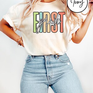 May include: Cream-colored t-shirt with the words "FIRST grade" in a colorful, block letter design. The word "grade" is in a cursive font. The shirt is paired with light blue denim jeans.