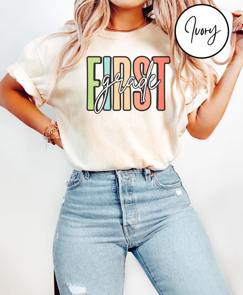May include: Cream-colored t-shirt with the words "FIRST grade" in a colorful, block letter design. The word "grade" is in a cursive font. The shirt is paired with light blue denim jeans.