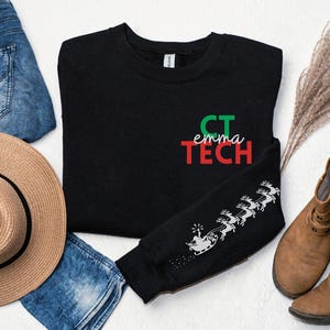 May include: Black sweatshirt with the text "CT emma TECH" in green and red. The sleeve features a white graphic of Santa Claus in a sleigh pulled by reindeer. The sweatshirt is displayed with a straw hat and blue jeans.