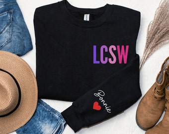 May include: Black sweatshirt with the text "LCSW" in a gradient of pink and purple on the front and the text "Bonnie" with a red heart on the left sleeve.