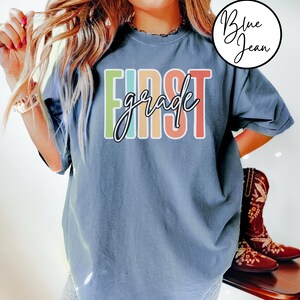 May include: A blue t-shirt with the words "FIRST grade" in a colorful, block letter design. The word "grade" is in a cursive font. The shirt is a relaxed fit and has short sleeves.