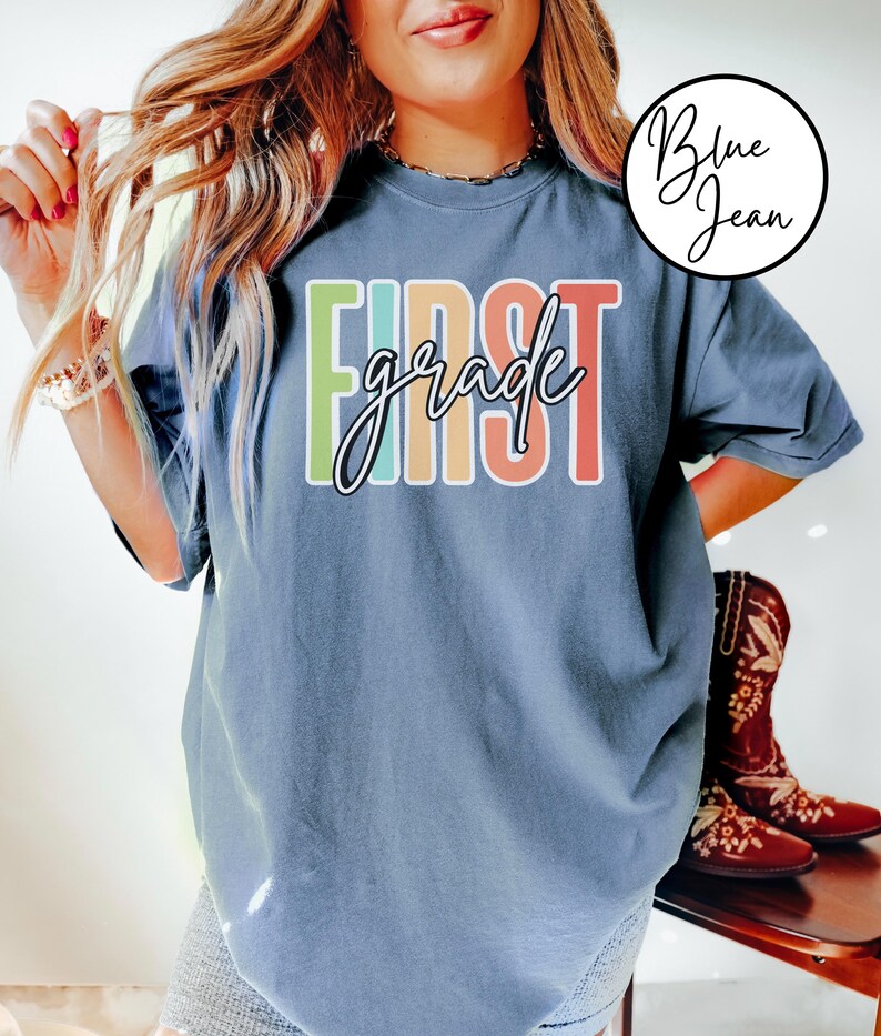 May include: A blue t-shirt with the words "FIRST grade" in a colorful, block letter design. The word "grade" is in a cursive font. The shirt is a relaxed fit and has short sleeves.