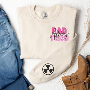 May include: A beige sweatshirt with the text "RAD Ashley TECH" in pink and black lettering. The sweatshirt has a black radiation symbol on the sleeve.