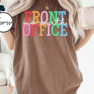 Pu&ograve; includere: Una maglietta marrone con il testo "Front Office" in colori arcobaleno. Il testo &egrave; in un font divertente e casual.