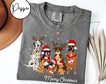 Puede incluir: Camiseta gris Comfort Colors con un diseño festivo de perros. El diseño incluye varios perros con gorros navideños, cornamentas y bufandas, con las palabras "Merry Christmas" en la parte inferior. Un collar plateado está drapeado sobre la camisa.