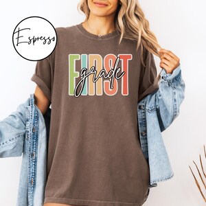 May include: A brown t-shirt with the words "FIRST grade" in a colorful, retro design. The shirt is worn with a denim jacket. A circular logo with the word "Espresso" is in the upper left corner.