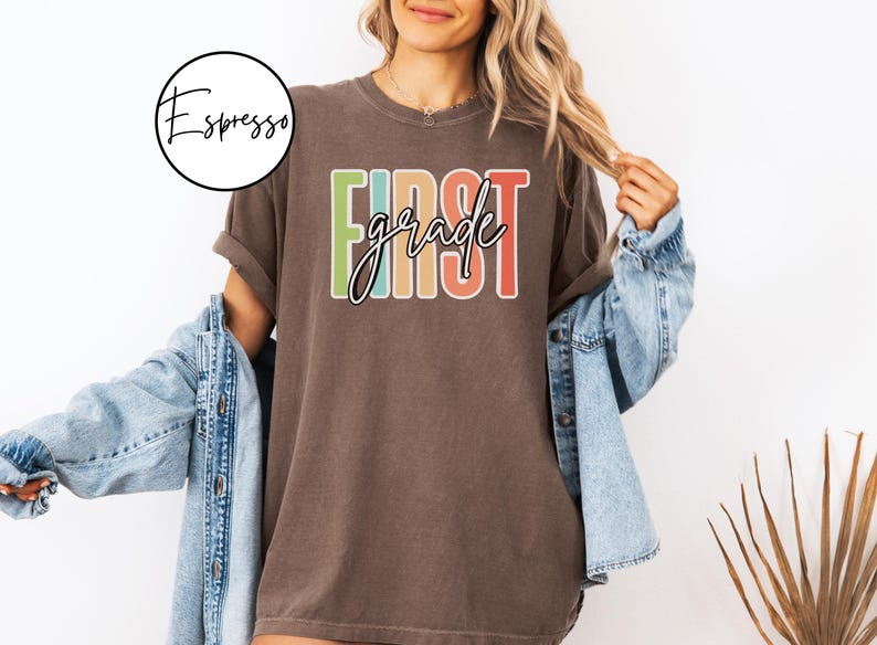 May include: A brown t-shirt with the words "FIRST grade" in a colorful, retro design. The shirt is worn with a denim jacket. A circular logo with the word "Espresso" is in the upper left corner.