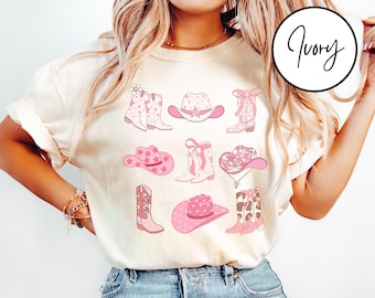 May include: A white t-shirt featuring a pink and white graphic design of cowboy boots and hats. The design includes a variety of cowboy boots and hats with stars and other patterns.