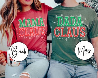 May include: Two Christmas-themed t-shirts. One is brick red with "MAMA CLAUS" in green and red, and the other is moss green with "DADA CLAUS" in green and red. Both shirts have snowflake accents.