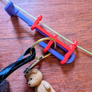 Può includere: Uno strumento in plastica viola e rossa con un filo sottile verde neon infilato, attaccato a un anello in metallo dorato. Una piccola statuetta di orso marrone pende dall'anello tramite una chiusura in metallo argentato, con un cinturino nero.
