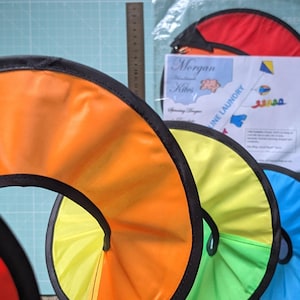 May include: A series of circular, collapsible fabric spinners in orange, yellow, green, blue, and purple, each with a black border. A ruler shows a 15-centimeter scale. A plastic bag contains a red spinner and a paper insert labeled Morgan Kites.