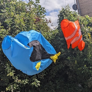 Puede incluir: Dos mangas de viento de tela con forma de pez colgadas al aire libre. Una es de color azul brillante con detalles en negro y amarillo, similar a un pez cirujano. La otra es de color naranja vibrante con rayas blancas, similar a un pez payaso.