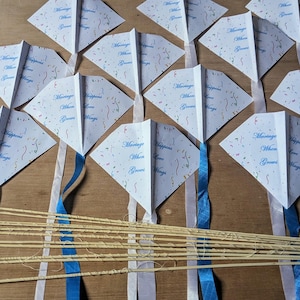 Puede incluir: Un conjunto de 12 cometas de papel de forma romboidal blanca con cintas azules y palos de bambú. Las cometas tienen un patrón de confeti y el texto "Marriage Happiness When Love Grows Wings".