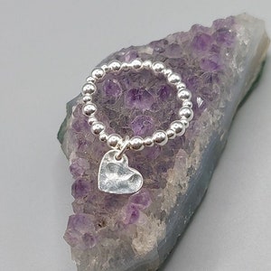 May include: A silver ring with a small, hammered heart charm hanging from it. The ring is made of small silver beads and is displayed on a bed of purple amethyst crystals.