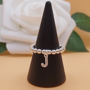 May include: A silver ring with a charm of the letter J hanging from it. The ring is made of small silver beads and is displayed on a black cone-shaped stand.