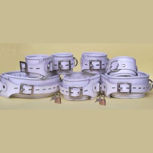 Puede incluir: Un conjunto de sujeciones de cuero blanco, que incluye un collar, esposas y tobilleras, cada una con hebillas plateadas y anillos metálicos. Se incluyen pequeños candados. Los artículos están dispuestos sobre un fondo amarillo claro.