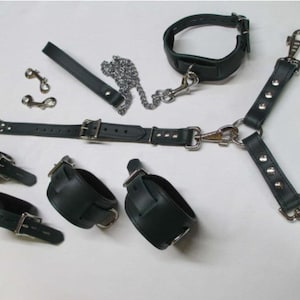 May include: A set of black leather restraints, including a collar, cuffs, and a harness. The set features silver-coloured metal hardware, such as buckles, clips, and a chain. The items are arranged on a white surface.