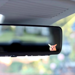 May include: A rearview mirror with a sticker of a cartoon character with a surprised expression. The character is a light pink color with a yellow outline.