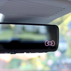 May include: A pink bow sticker on a rearview mirror in a car.