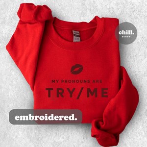 May include: Red sweatshirt with the text "MY PRONOUNS ARE TRY/ME" in black lettering. A red lip print is above the text. The text "embroidered." is below the text on the sweatshirt.