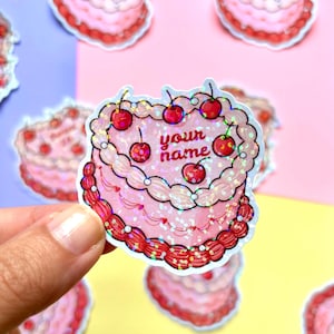 May include: A pink cake sticker with a cherry design and the text "your name". The sticker has a glittery, holographic effect. Other similar stickers are in the background.