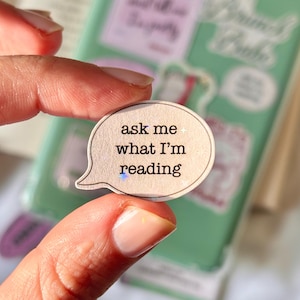 May include: A speech bubble shaped sticker with a light beige background and a thin dark outline. The text printed in black, sans-serif font reads: ask me + what I'm reading. The sticker has a subtle holographic finish that catches the light.