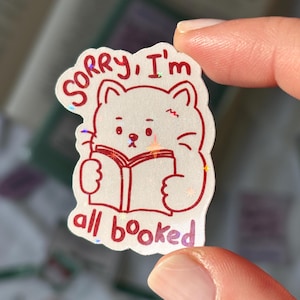 Peut inclure: Autocollant de chat blanc avec un contour rouge et le texte "Sorry, I'm all booked." Le chat tient un livre. L'autocollant a un effet holographique et scintillant.