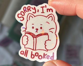 Op de afbeelding: Een witte kattensticker met een rode omtrek en de tekst "Sorry, I'm all booked." De kat houdt een boek vast. De sticker heeft een holografisch, glinsterend effect.
