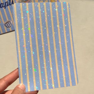Könnte beinhalten: Eine rechteckige Karte mit einem vertikalen Streifenmuster in Hellblau und Beige. Die Oberfläche der Karte ist mit einem holografischen Finish versehen, das kleine, mehrfarbige sternförmige Glitzerpunkte über das gesamte Streifendesign zeigt.