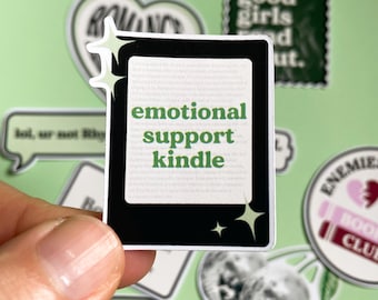 Op de afbeelding: Een rechthoekige sticker met een zwarte e-reader met een wit scherm waarop de groene tekst 'emotional support kindle' en kleine stervormige accenten staan. De sticker heeft een witte rand en wordt tegen een lichtgroene achtergrond gehouden.