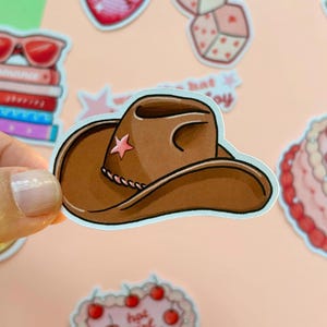 May include: A brown cowboy hat sticker with a pink star. The hat has a curved brim and a decorative rope detail around the crown. Other stickers with a romance theme are in the background.