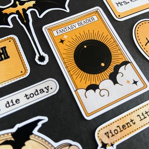 Peut inclure: Un ensemble d'autocollants comprenant une carte rectangulaire avec le texte FANTASY READER, un soleil noir et des nuages. D'autres autocollants incluent une silhouette de dragon noir, un badge Hi, my name is Mrs. Riorson, et les textes die today et Violent lit.