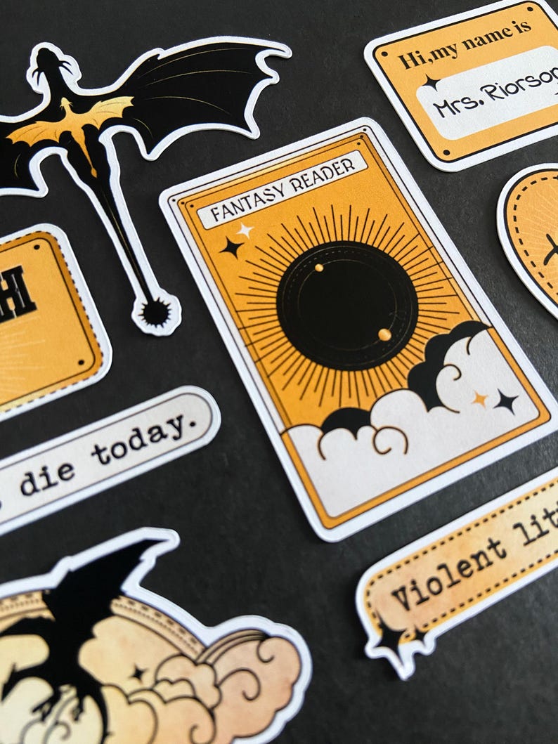 Peut inclure: Un ensemble d'autocollants comprenant une carte rectangulaire avec le texte FANTASY READER, un soleil noir et des nuages. D'autres autocollants incluent une silhouette de dragon noir, un badge Hi, my name is Mrs. Riorson, et les textes die today et Violent lit.