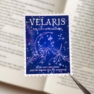 Könnte beinhalten: Ein rechteckiger Briefmarkenaufkleber mit einem tiefblauen Aquarell-Nachthimmel, einer weißen Mondsichel, Berggipfeln und Sternen. Der Text lautet VELARIS THE CITY OF STARLIGHT und to the stars who listen and the dreams that are answered.