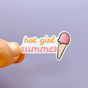 May include: A die-cut sticker featuring the text hot girl summer in orange and pink cursive lettering, accompanied by a graphic of a pink ice cream scoop on a tan waffle cone, all outlined in white against a solid light purple background.