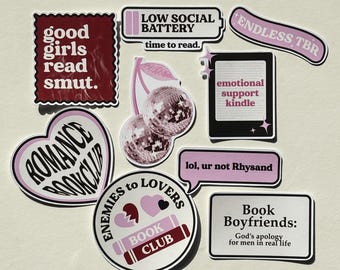 May include: A collection of vinyl stickers featuring book-themed designs in shades of pink, maroon, and black. Text includes "good girls read smut," "LOW SOCIAL BATTERY," "ENDLESS TBR," "emotional support kindle," "lol, ur not Rhysand," and "Book Boyfriends."