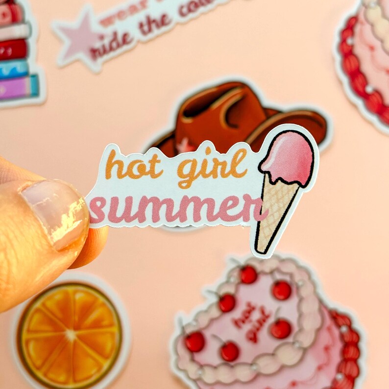 May include: A die-cut sticker featuring the text "hot girl summer" in orange and pink script lettering, accompanied by a graphic of a pink ice cream cone with a waffle texture. The sticker has a white border and is held against a light background.